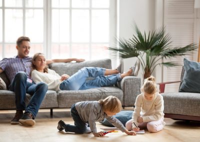 Children sister and brother playing drawing together on floor while young parents relaxing at home on sofa, little boy girl having fun, friendship between siblings, family leisure time in living room